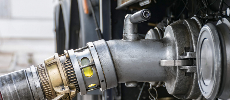 A close-up image of a fuel nozzle attached to a pump, likely at a gas station, with mechanical components of the pump visible in the background.