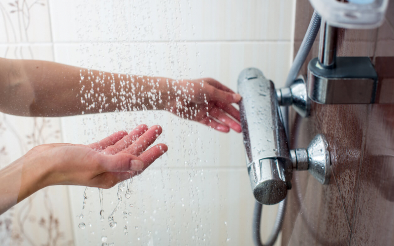 A person is adjusting a handheld shower head spraying water, with focus on the water droplets and the person's hands against a tiled background.